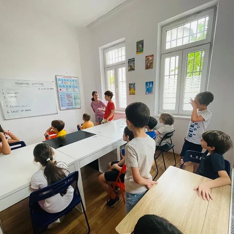 Copii la mesele dintr-o sală luminoasă, în timpul unei activități în limba engleză scrise pe tabla albă