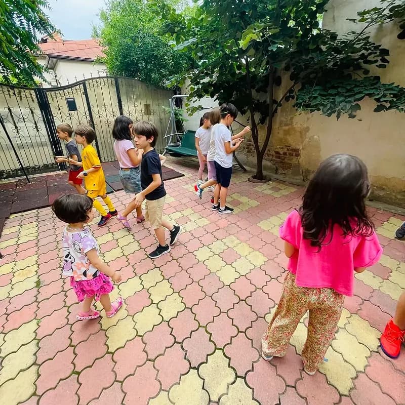 Copii alergând și jucându-se în curtea pavată a after school-ului, la umbra copacilor