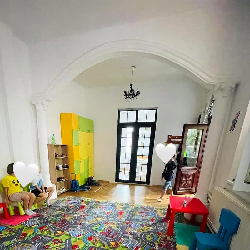 Sala de joacă luminoasă, cu covor cu străzi, scăunele colorate, dulapuri galben cu verde și ușă mare cu geam